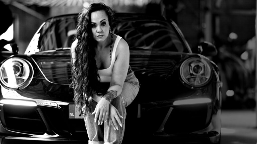 Lana Lain sitting confidently in front of a black sports car in a bold black-and-white portrait — powerful, feminine, and rebellious.